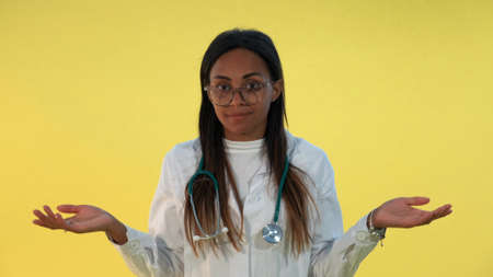 Close-up of mixed-race female doctor spreading hands to the sides on yellow background. She is lab coat and with stethoscope on shoulders.の写真素材