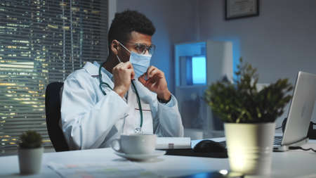 Medium shot of Young african doctor showing how to wear medical mask by video call on computer. He working home because of pandemic.の写真素材