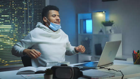 Tired multiethnic man with protective mask working home late at night and making warm up activities. City in lights in the background.の写真素材