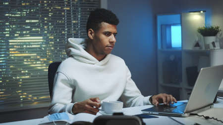 Handsome multiracial man drinking a cup of coffee and working on computer at night. There are skyscrapers in the background.の写真素材