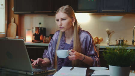 Working at home: Busy business woman in home bathrobe drinking a cup of coffee. She sitting at the computer in the kitchen.の写真素材