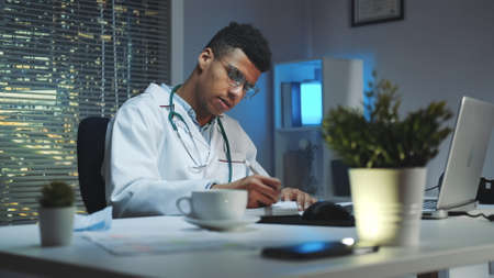 Mixed-race doctor making notes and talking by video call on computer. He working home because of COVID-19 pandemic.の写真素材