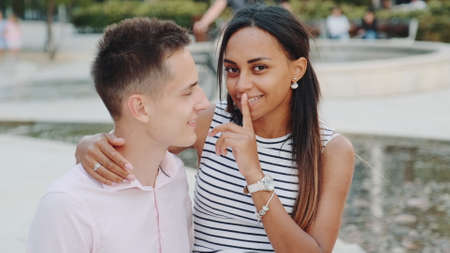 Pretty black lady whispering something in boyfriends ear and then showing keep silence gesture to the camera with point finger and mouth. She telling him some special things.の写真素材