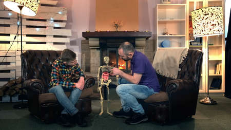 Father explaining his elder son the bone structure of human body showing on skeleton. They sitting on comfortable armchairs near the fireplaceの写真素材
