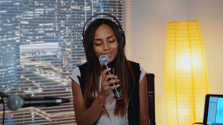 Close-up of beautiful multiracial girl in headphone singing into microphone and recording a song in home studio with skyscrapers in the background.の写真素材