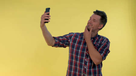 Attractive tall boy making selfie on smartphone. He smiling and standing against yellow background.の写真素材
