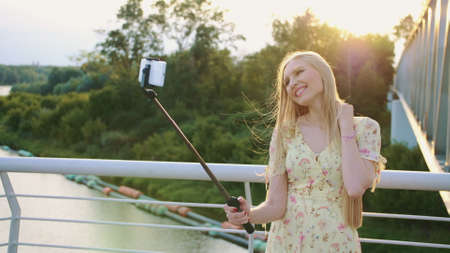 Woman taking selfie on bridge. Cheerful pretty blonde woman standing on pedestrian bridge and taking selfie with monopod in sunset.の写真素材