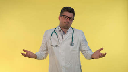 Good-looking young doctor in lab coat showing that he cant help in this situation and spreading hands to the sides.. Yellow background.の写真素材