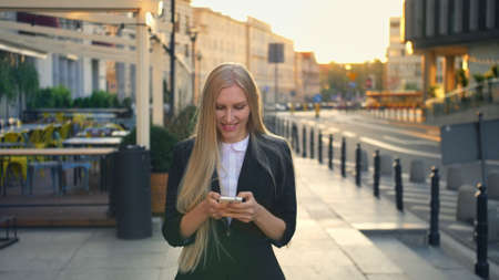 Elegant blond woman in suit walking on street and browsing smartphone with smile against urban background.の写真素材