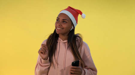 Happy african woman in Santa hat and wireless headphones listening to the music on yellow background. She singing some Christmas songs.の写真素材