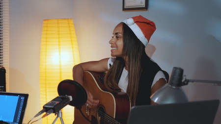 Beautiful black woman in Santa hat singing and playing guitar on Christmas Eve in cozy home studio. In front there is a recording equipment.の写真素材