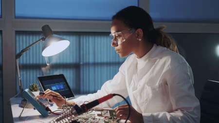 Close up of female engineer of electronics in white coat and protective glasses checking motherboard with multimeter tester and other electronic devisesの写真素材