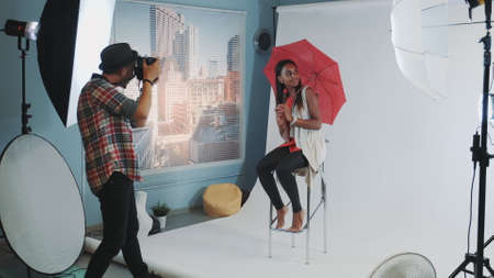 Stylish african model posing with red umbrella on bar high chair for fashion magazine photo shoot. Photographer taking photos on professional camera.の写真素材