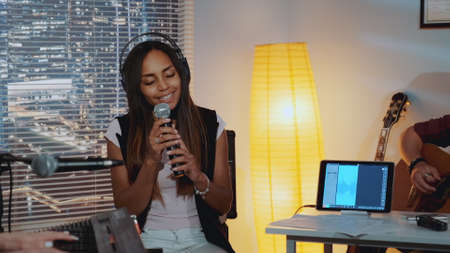 Attractive black woman with close eyes emotionally singing song into microphone and gesturing while her band playing the instruments in home studioの写真素材