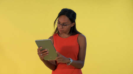 Black business woman in dress and eyeglasses analysing something on tablet There is yellow background.の写真素材