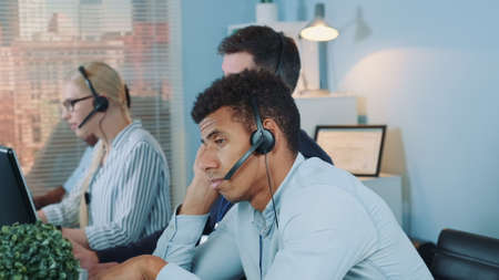 Bored multiethnic customer support operator waiting for telephone call from client. His colleagues are busy working.の写真素材