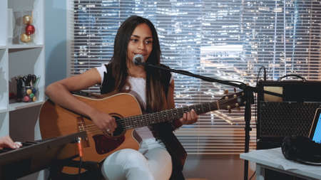 Black woman vocalist recording a song and playing guitar in home studio. There are skyscrapers in the background.の写真素材