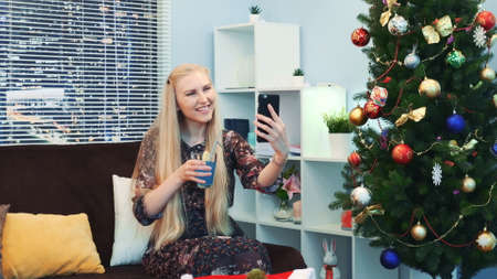 Beautiful young lady is making video call by smartphone. Shes sitting on sofa with a glass of drink in the living room decorated with Christmas tree and lights with skyscrapers in the background.の写真素材