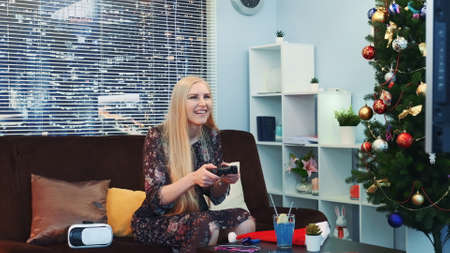 Medium close up of nice girl playing on joystick near the Christmas tree. There are also VR glasses headset on sofa in the room with skyscrapers in the background.の写真素材