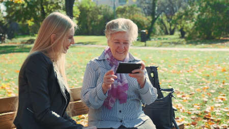 Elder grandmother showing her granddaughter photos on smartphone. They sitting on bench and talking in autumn park.の写真素材
