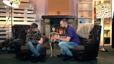 Father explaining his elder son the bone structure of human body showing on skeleton. They sitting on comfortable armchairs near the fireplaceの写真素材