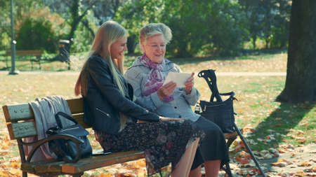 Old woman showing her pretty granddaughter a letter. They sitting on a bench in park one autumn day.の写真素材
