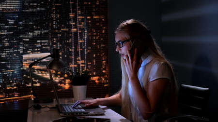 Young female manager working on the computer and speaking with somebody by smartphone in her cosy office late at nightの写真素材