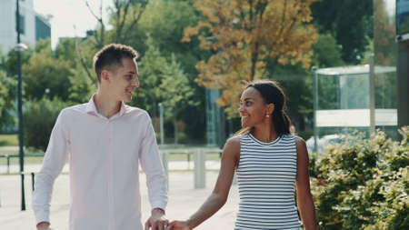 Multi-ethnic couple shyly looking at each other while walking the street, holding hands and talking. Relationship concept.の写真素材