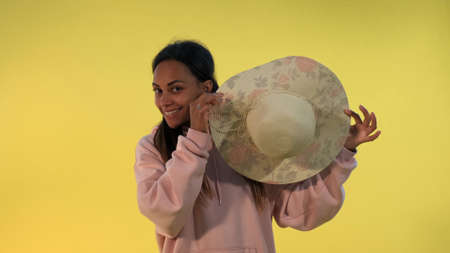 Smiling african woman hiding behind the hat on yellow background. She playing with someone.の写真素材