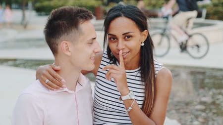 Pretty black lady whispering something in boyfriends ear and then showing keep silence gesture to the camera with point finger and mouth. She telling him some special things.の写真素材