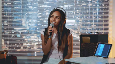 Stylish mixed-race girl singing into microphone at the rehearsal in home studio with skyscrapers in the background.の写真素材