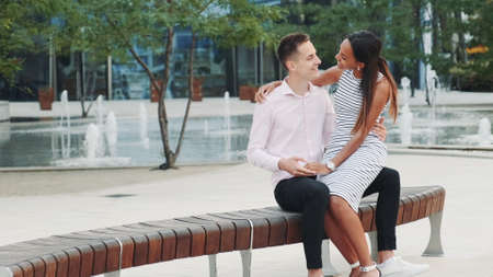 Cheerful multiracial couple having date outdoors near the fountains. They sitting on the bench, hugging, kissing and looking at each other.の写真素材