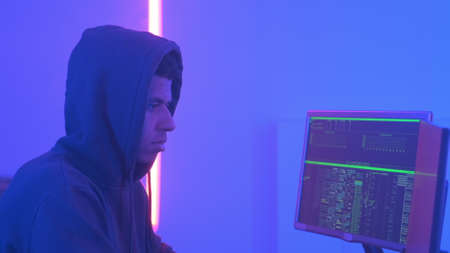 Profile view of african hacker in hoodie working in room filled with display screens and warm colored neon lights.の写真素材