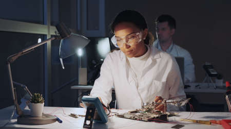 African american electronics engineers checking electronic board with multimeter tester and other electronic devises in lab. There is a fellow worker in the background.の写真素材