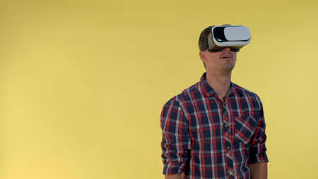 Dark-haired man using virtual reality glasses while standing against yellow background. He is in checkered shirt.の写真素材