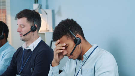 Medium shot of Bored and dissatisfied black call center agent talking to the client on phone. He is behaving nervously. He working with multiracial young team.の写真素材