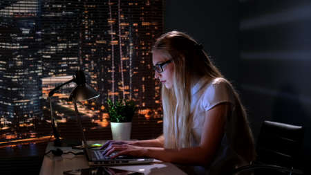 Pretty psychologist searching books in Internet in her office late at night. In the background there are skyscrapers.の写真素材