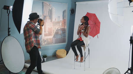 Stylish african model posing with red umbrella on bar high chair for fashion magazine photo shoot. Photographer taking photos on professional camera.の写真素材