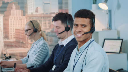 Close-up shot of smiling mixed-race man taking call in busy call center. In the background there are his colleagues also talking to clients on phone.の写真素材