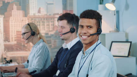 Close-up shot of smiling mixed-race man taking call in busy call center. In the background there are his colleagues also talking to clients on phone.の写真素材