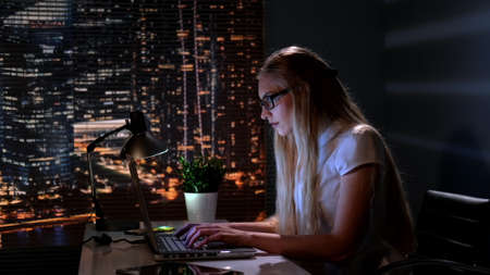 Pretty psychologist searching books in Internet in her office late at night. In the background there are skyscrapers.の写真素材