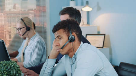 Bored multiethnic customer support operator waiting for telephone call from client. His colleagues are busy working.の写真素材