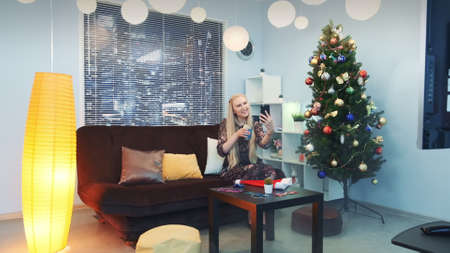 Young woman is making video call by smartphone with skyscrapers in the background. Shes sitting on sofa with a glass of drink in the living room decorated with christmas tree and lights among smoke.の写真素材