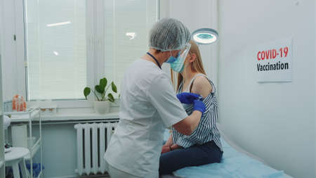 Female doctor in medical protective cloths injecting in womans arm a vaccine against coronavirus. Covid-19 vaccination concept.の写真素材