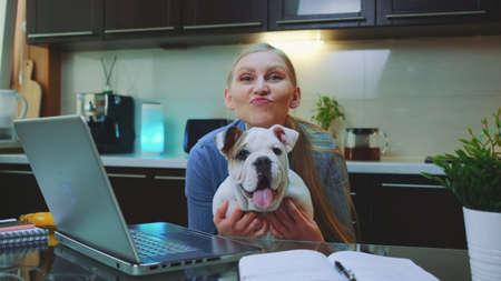 Happy woman with a bulldog looking to the camera and smiling. They sitting at the table with computer in the kitchen.の写真素材