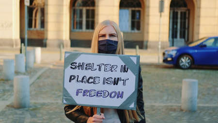 Woman in protective mask protesting that shelter in place does not mean freedom. Young blonde woman walking down the street with placard.の写真素材