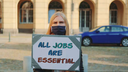 On protest march woman in protective mask drawing attention to the importance of all kind of jobs. She holding banner and walking on the street.の写真素材