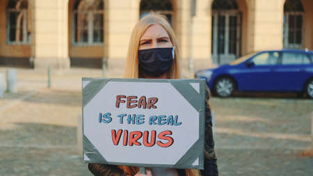 Blonde woman in protective mask protesting that fear is the real virus on protest march. She holding banner in her hands and walking on the street.の写真素材