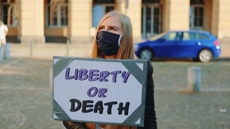 Blonde woman in protective mask with protest banner calling to choose liberty or death. She walking on the street.の写真素材