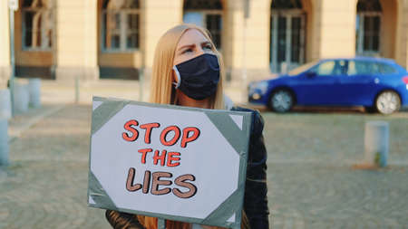 Concerned woman in protective mask with protest banner calling to stop lies by walking on the city streetの写真素材
